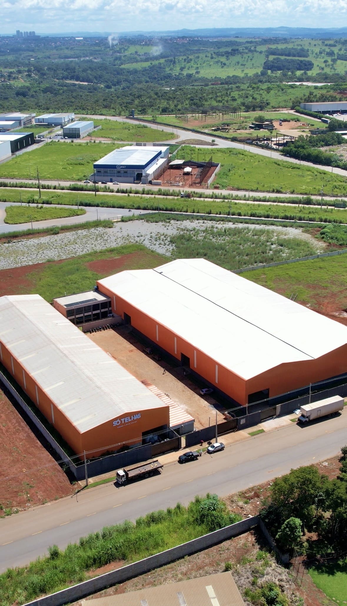 Vista aérea da fábrica Só Telhas em Aparecida de Goiânia, galpões industriais laranja e branco com pátio de manobra, caminhões de entrega estacionados, paisagem verde ao redor, iluminação natural de dia claro, fotografia profissional ultra-realista